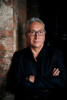 Distinguished looking gentleman wearing dark-rimmed glasses leaning against the corner of a brick wall