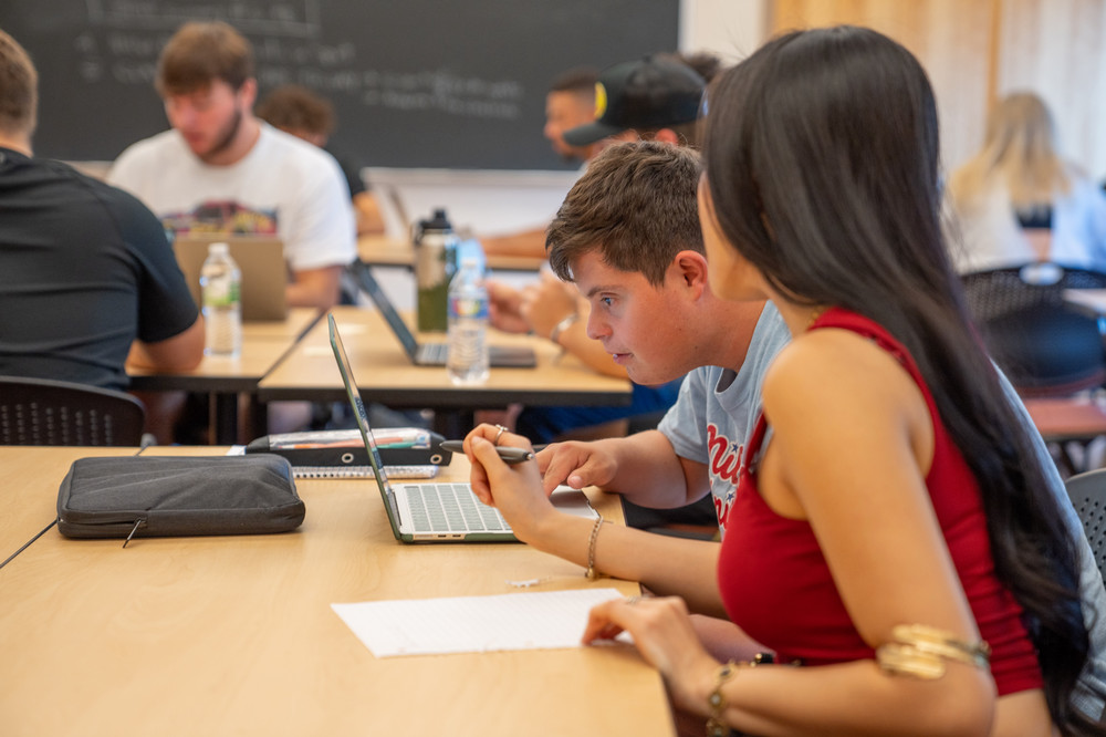 A male student sits in front of a laptop. A female student sits next to him and points at his screen.