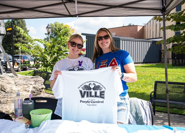 Two women wearing sunglasses stand under a canopy tent at an outdoor campus event, smiling and holding up a white T-shirt that reads “VILLE Purple Carnival.” 