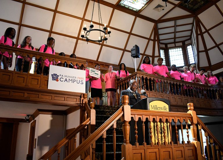 Dr. Wubah stands at a podium on a staircase inside a wood-paneled hall at Millersville, addressing a crowd. Students wearing bright pink shirts line the balcony above, looking down. A banner reading “PA Hunger-Free Campus” hangs from the railing, and a chandelier and skylight are visible overhead.