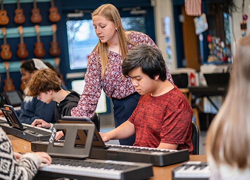 A music teacher stands over a male student, guiding him as he plays a digital keyboard in a classroom filled with other students practicing on their own key