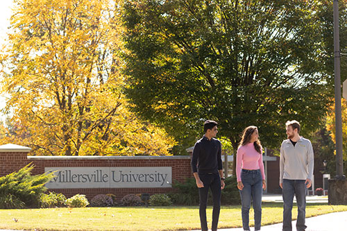 Three students walk past a brick sign that reads Millersville University on a sunny autumn day.