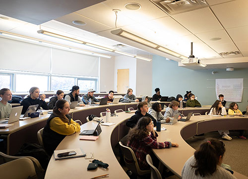 A classroom setting with students seated along curved tiered desks facing towards the right side of the room, many with laptops open.