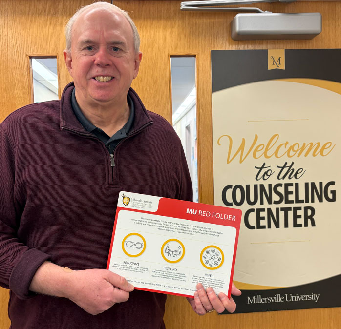 Dr. Lynch holding a red folder standing in front of a door with a sign welcoming students to the counseling center.