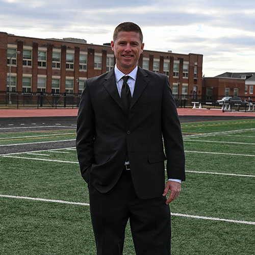 Man in a suit sanding on Biemesderfer field with an academic building in the background