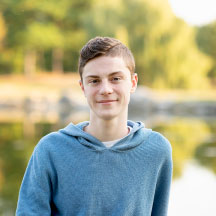Person standing outdoors near a lake with trees in the background.