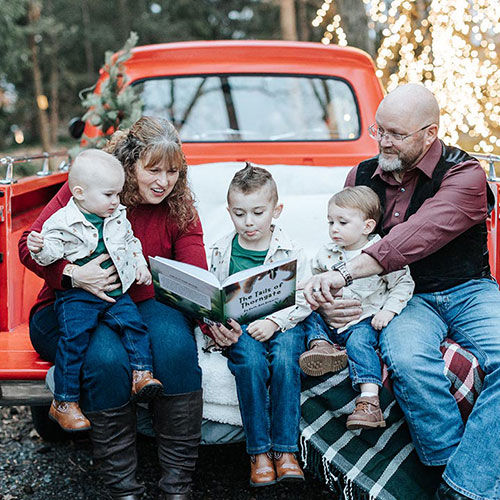 Family sitting on the tailgate of a red pickup truck reading a book together in a festive outdoor setting.
