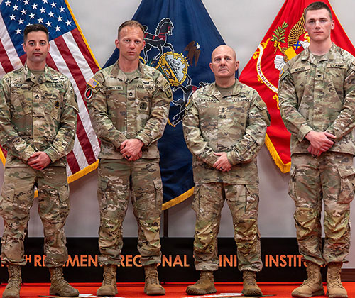 Four men in camouflage uniforms stand at attention in front of three flags.
