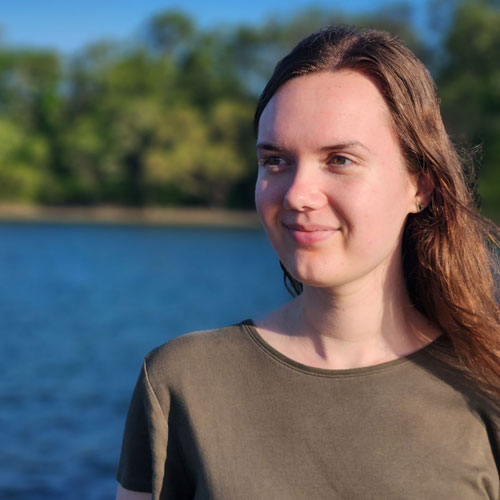 Person in olive green shirt standing near a lake with trees.