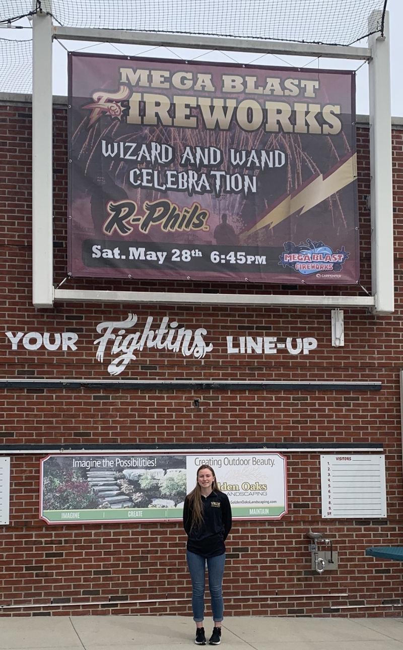 Ashly standing below her printed banner for her internship with Reading Fightin Phils.